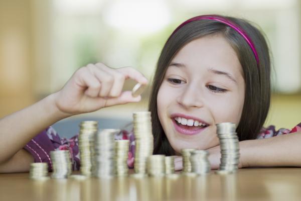 A young child with long hair rests their forearms on a table with piles of different coins. The piles are different sizes, and the child is smiling while placing a coin at the top of the highest pile.